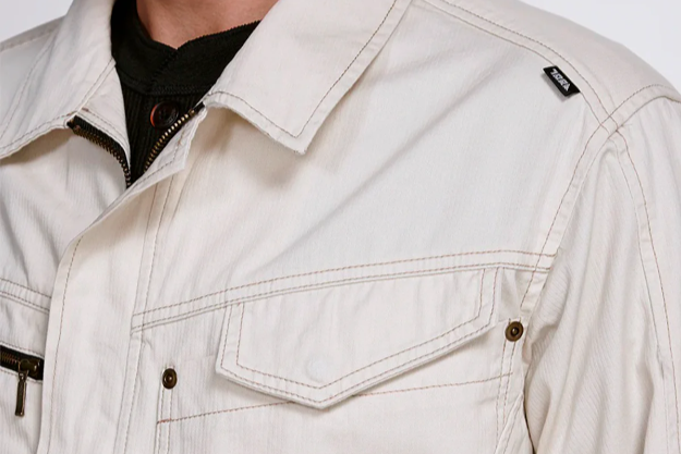 A close-up of an off-white work jacket with contrast stitching, a pointed collar, and a flap chest pocket, worn over a black inner shirt.