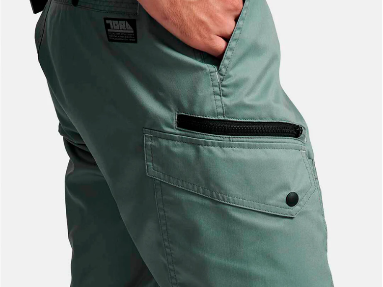A close-up of light green work pants featuring a zippered side pocket, flap pocket with snap button, and a hand tucked into the front pocket.