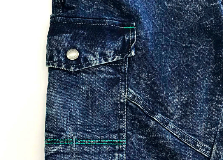 A close-up of dark blue denim work pants featuring a snap-button flap pocket, contrast stitching, and a curved seam detail.