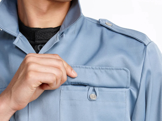 A close-up of a light blue work jacket with a stand collar and flap chest pocket, with the model lifting the pocket flap by hand.