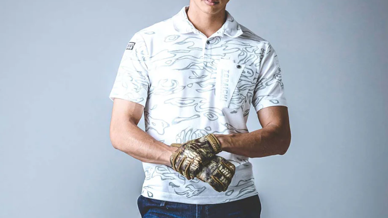 A model wearing a white short-sleeve polo shirt with a light gray abstract pattern and chest pocket, standing with gloved hands against a soft blue-gray background.
