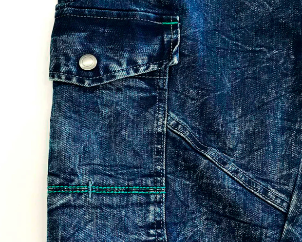 Close-up of dark indigo denim workwear fabric with a flap pocket, metal button, green stitching, and visible seam details.